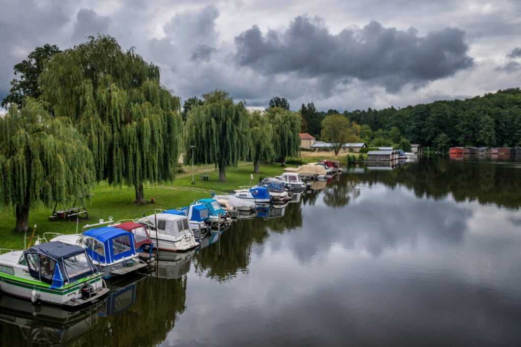 A serene view of a marina with boats, lush weeping willows, and cloudy skies.